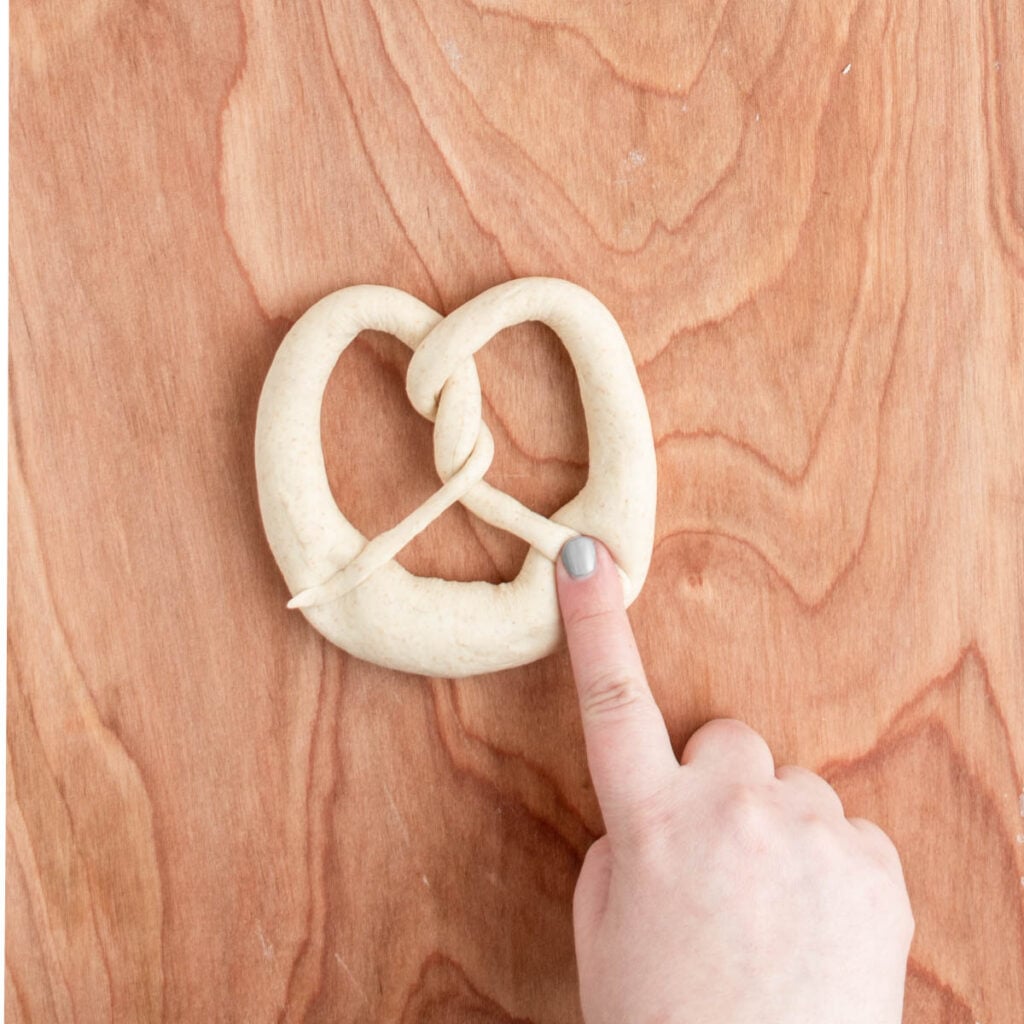 A hand with light skin and gray nail polish points to a piece of raw sourdough beer pretzel dough shaped into a knot on a wooden surface.