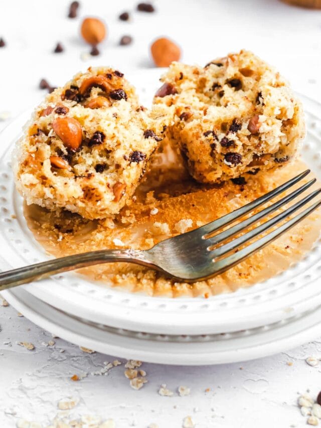 a muffin that's been split open on a small plate with a fork beside it. the inside is filled with chocolate chips and large hazelnut chunks.
