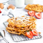 A stack of 3 funnel cakes on a small cooling rack. two halves of a strawberry sit in front of them.