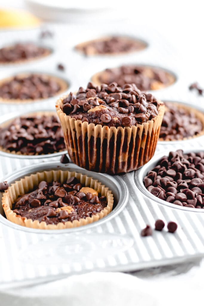 a chocolate muffin sits on top of a muffin tin with other muffins in the wells around it