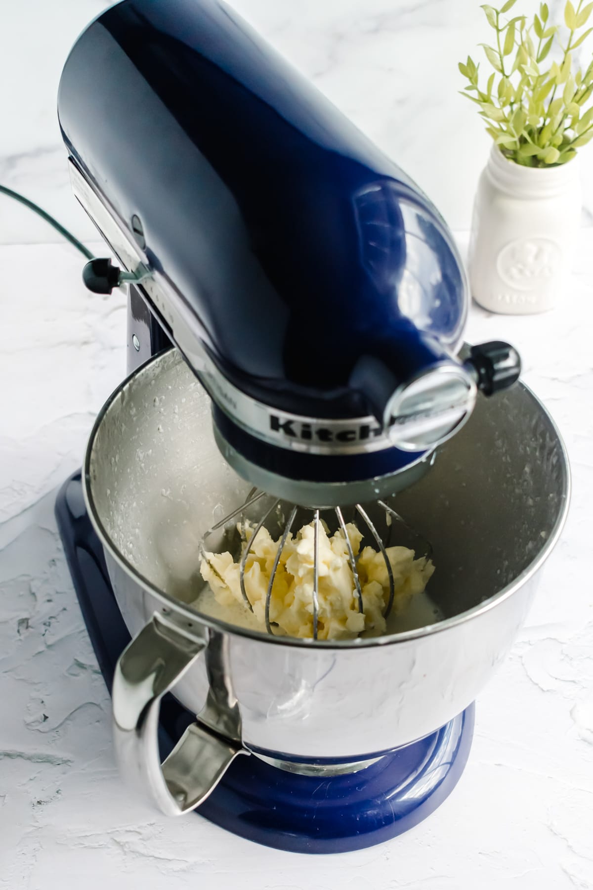 A blue stand mixer with a metal bowl with butter collected inside the whisk attachment. A small potted plant sits in the background.