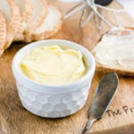 a small white ramekin of homemade butter on a wooden board with a butter knife and slices of bread
