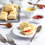 a plate of mini honey butter biscuits with jam sits in front of a cooling rack with other mini biscuits and bowls of honey butter and jam