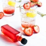 a small bottle of strawberry syrup lies next to a strawberry soda with lemon and strawberry garnish\