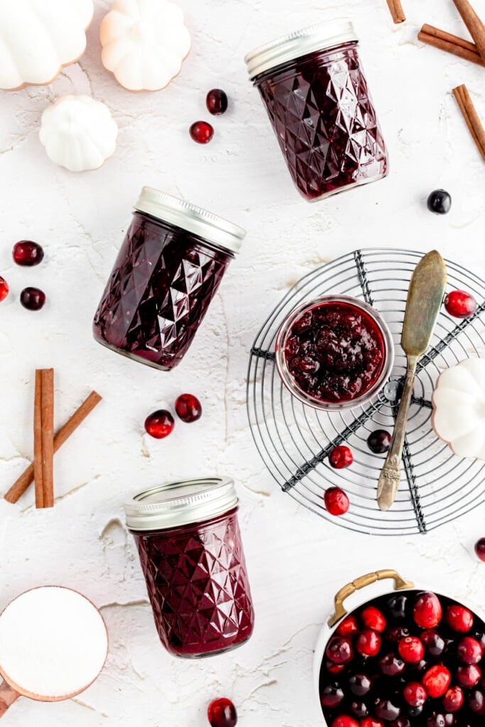 Three jars of spiced cranberry jam with silver lids, a small bowl of cranberry sauce with a spoon, loose cranberries, cinnamon sticks, white pumpkins, and a pot of cranberries arranged on a white surface.
