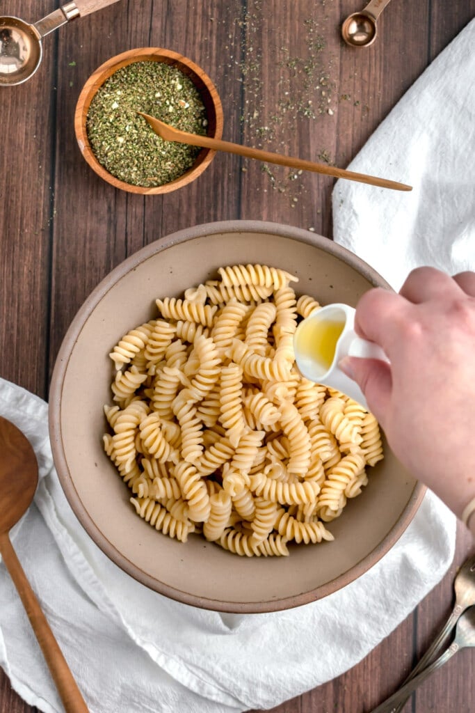 A hand pours oil over a bowl of cooked spiral pasta on a wooden table, preparing it for a delicious pesto with dried basil. A small bowl of green herbs and kitchen utensils are nearby on a white cloth.