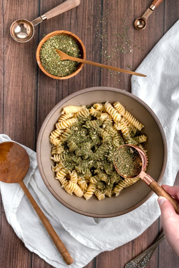 A hand sprinkles dried basil pesto from a copper measuring cup onto a bowl of cooked pasta, creating the perfect base for a quick pesto with dried basil. A bowl of herbs, measuring spoons, a wooden spoon, and a white cloth are on a wooden table nearby.