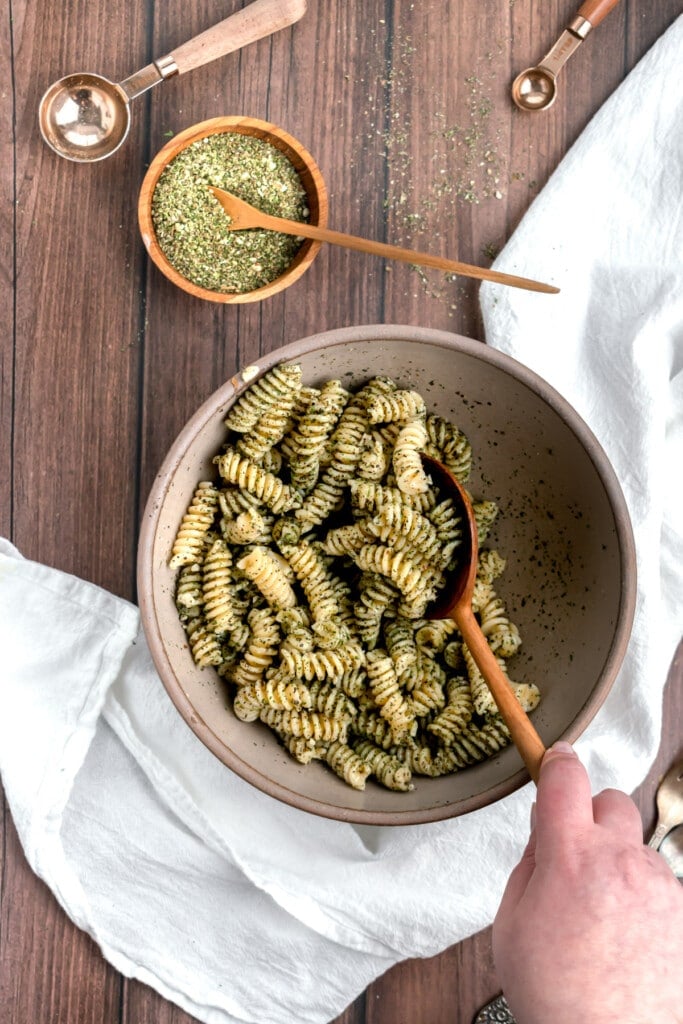 A hand stirs a bowl of spiral pasta coated with earthy green pesto made with dried basil. A wooden spoon is used. A small bowl of herbs, measuring spoons, and a white cloth are arranged on a wooden surface.