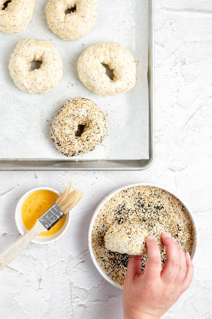 A hand dips an unbaked everything bagel into a bowl of mixed everything bagel topping. Other unbaked bagels rest on a parchment-lined baking tray, with a small bowl of egg wash and a pastry brush nearby on a white surface.