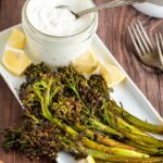 close up of cumin roasted broccolini on a platter with lemon wedges and a jar of lemon yogurt dipping sauce