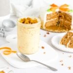 a jar of carrot cake ice cream topped with crumbled carrot cake. a spoon is on the white tray in front of the jar. a sliced carrot cake is behind it to the right.