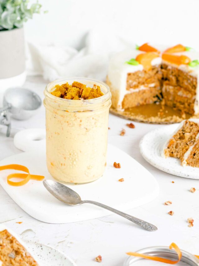 a jar of carrot cake ice cream topped with crumbled carrot cake. a spoon is on the white tray in front of the jar. a sliced carrot cake is behind it to the right.