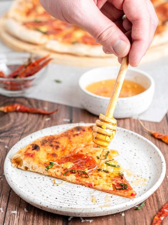 a hand drizzles honey on a pizza slice with a honey wand.