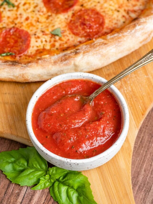 a small bowl of red pizza sauce on a pizza peel with a pepperoni pizza behind it.