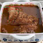 a braised, thinly sliced brisket in the braising liquid in a white casserole dish on a tan napkin.