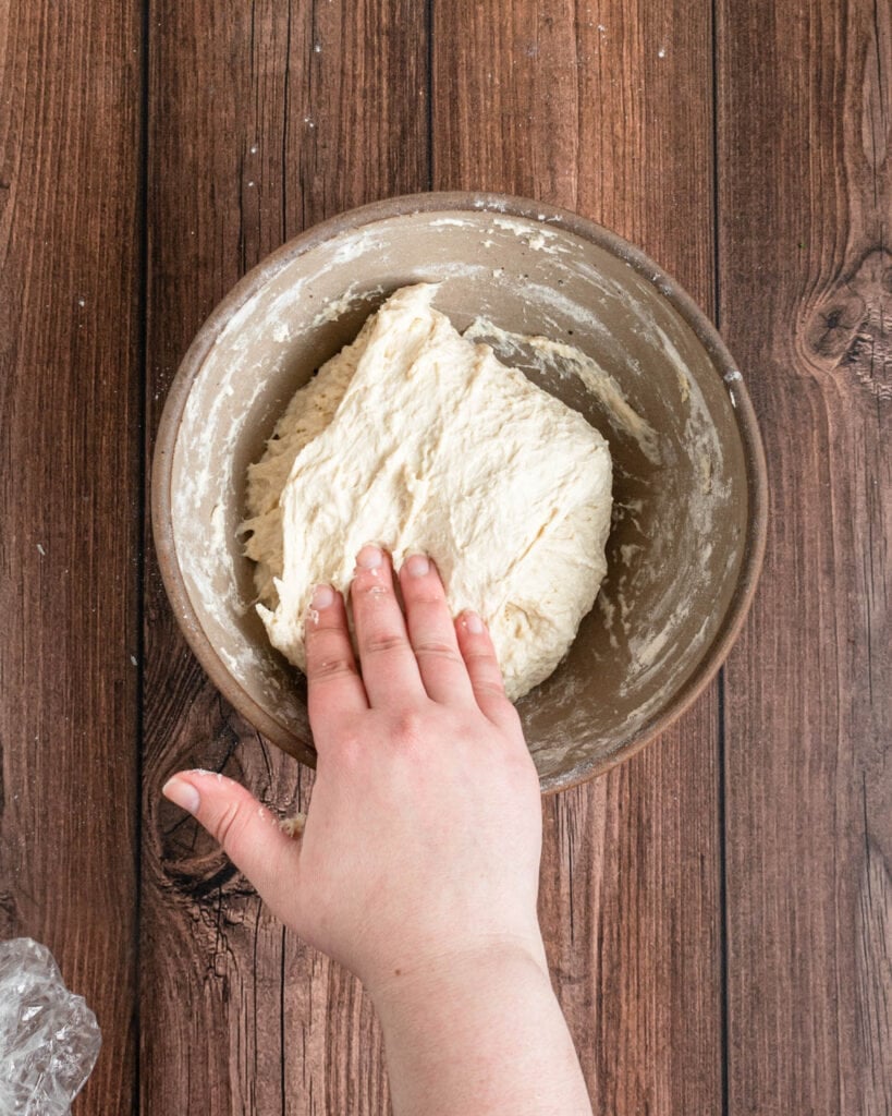 A hand folds down the top edge of a ball of focaccia pizza dough in a brown mixing bowl, which rests on a wooden surface. Some flour is visible on the dough and bowl.