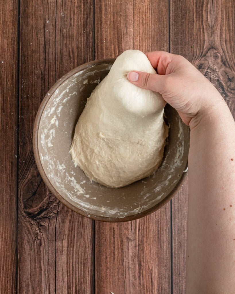 A hand stretches the top edge of a focaccia dough upwards from a brown bowl on a wooden surface, showing the elasticity perfect for making focaccia pizza.