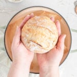two hands holding a small grapefruit sized loaf of crusty bread over a dutch oven.
