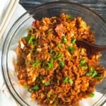 an overhead shot of a large glass bowl with breakfast fried rice in it. a wooden spoon sticks out of the rice and the bowl sits on an off-white linen napkin on a grey wooden counter. a pair of chopsticks sits to the left of the bowl.