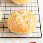 brioche bun topped with sesame seeds and an X cut into it sits on a black wire cooling rack.