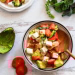an overhead shot of a chopped caprese salad in a bowl with a wooden spoon sticking out of it. on the wooden table next to it are two tomato slices, an empty avocado skin, and some fresh basil leaves.