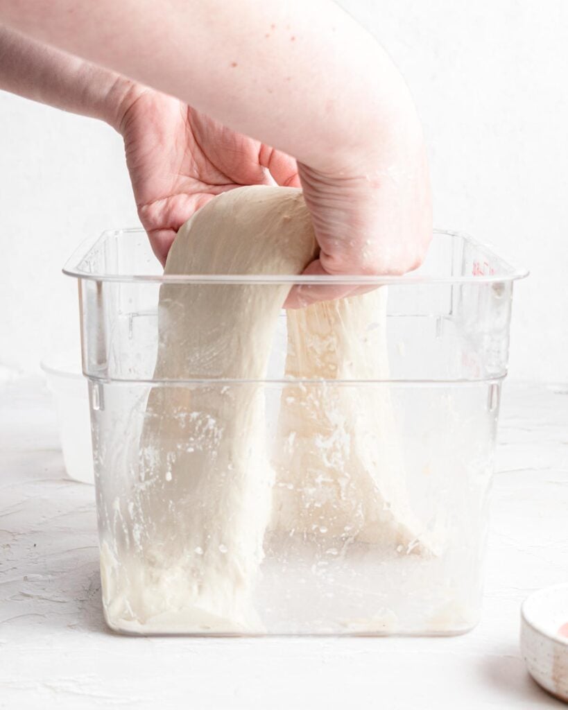 A person performs a coil fold technique with ciabatta bread dough inside a clear plastic container on a white surface.