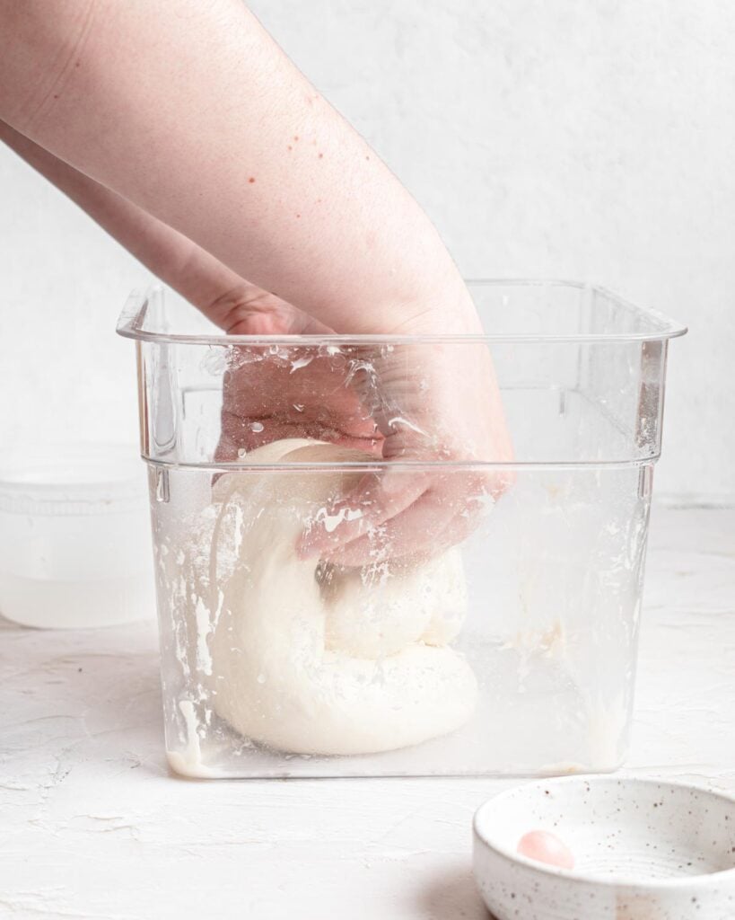 A hand is kneading ciabatta bread dough in a clear plastic container. The container has traces of flour on the sides, and a small bowl with a pink object sits on the nearby white surface.
