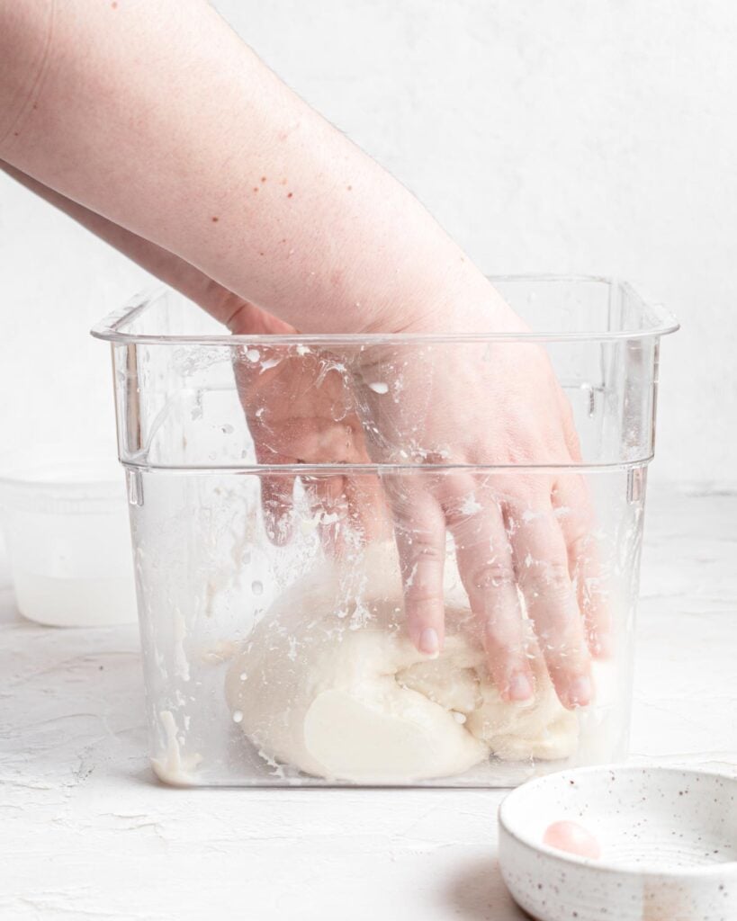 A hand is kneading or mixing ciabatta bread dough inside a clear rectangular container on a white surface, with a small bowl and another container nearby.