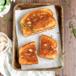 two grilled cheese sandwiches on a tarnished metal sheet pan lined with parchment paper. one has been cut in half. there are sprigs of thyme decorating the scene.