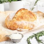 a loaf of rosemary parmesan bread on a sheet of parchment paper with a small cup of parmesan cheese and fresh rosemary sprigs nearby.