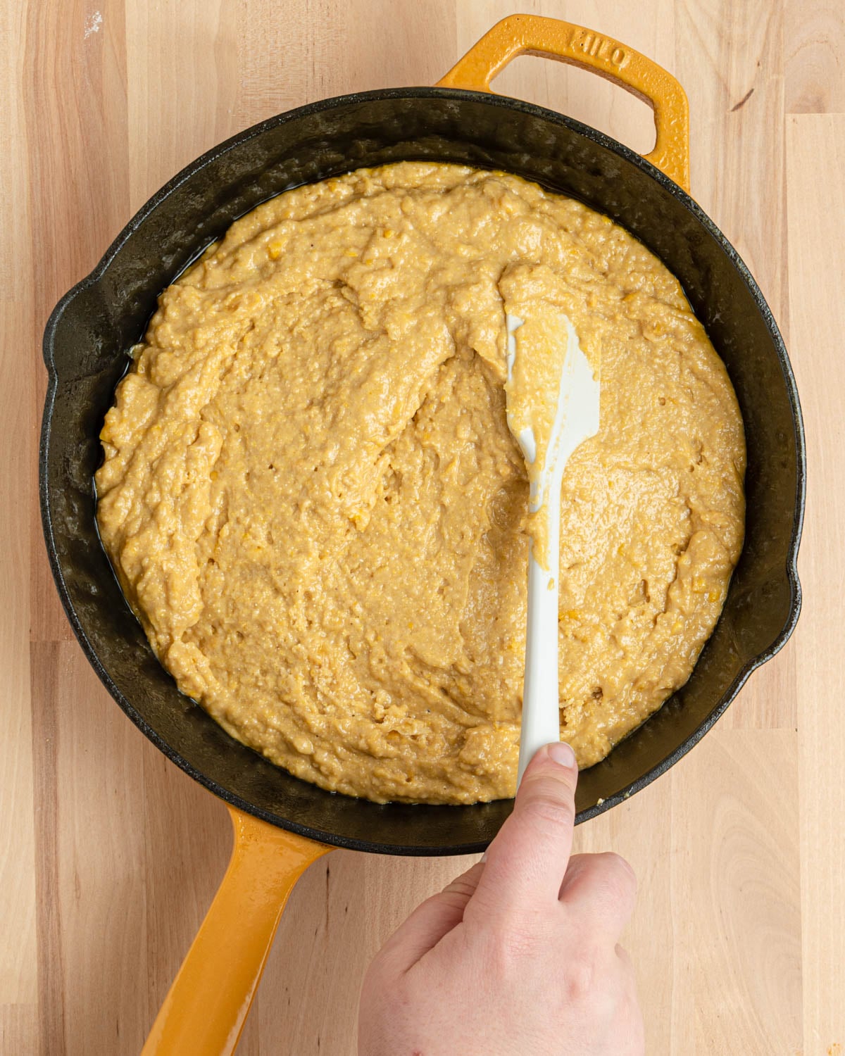 Using a narrow white spatula to smooth the cornbread batter into a cast iron skillet.
