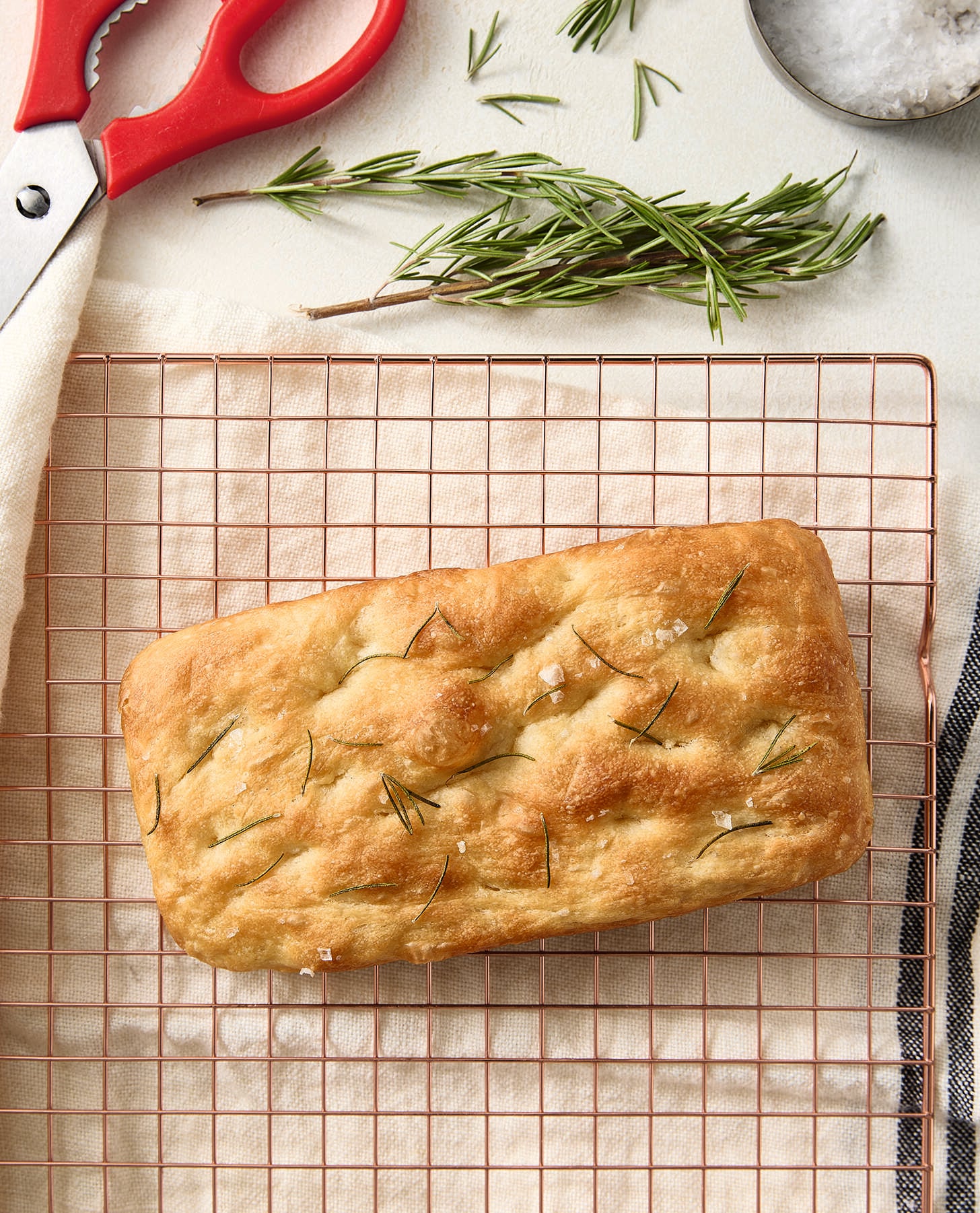 A rectangular loaf of focaccia bread with rosemary sits on a cooling rack. Fresh rosemary, a red-handled pair of scissors, and a bowl of salt are placed nearby on a light surface.