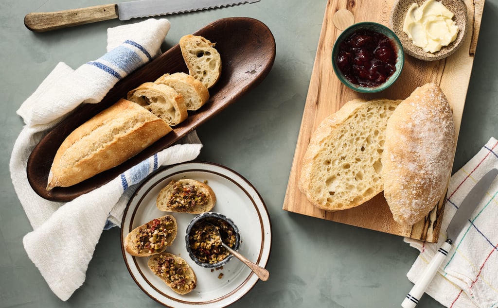 A table with a wooden tray holding a baguette, a sliced roll on a board with butter and jam, and a plate of sliced bread topped with a chopped nut and olive mixture. Knives and a striped towel are nearby.