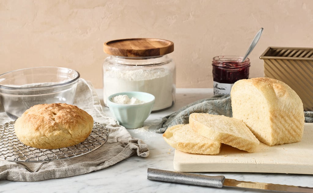 Round and sliced loaves of bread on a cooling rack and cutting board, with a jar of flour, a small bowl of flour, a jar of jam with a spoon, a loaf pan, and a bread knife on a marble countertop.