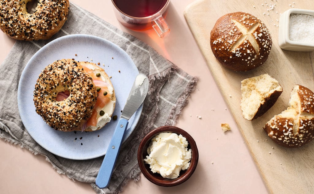 A plate with two bagels, one topped with smoked salmon and cream cheese, sits next to a cup of tea, a bowl of cream cheese with a knife, and two pretzel buns on a cutting board with a container of coarse salt.