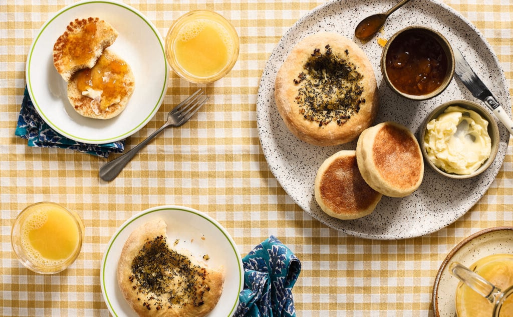 A table set with English muffins, butter, marmalade, and two glasses of orange juice on a yellow checkered tablecloth. Some muffins are split with spreads, and blue-patterned napkins are beside the plates.