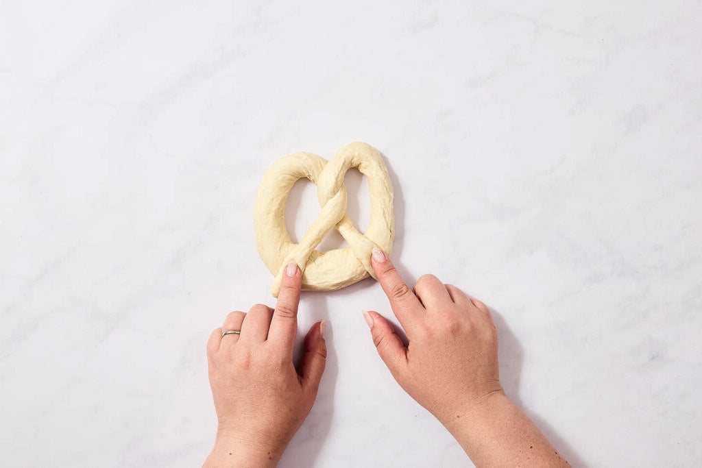Two hands shape a piece of dough into a pretzel on a white surface. The person’s left hand wears a gold ring on the ring finger.