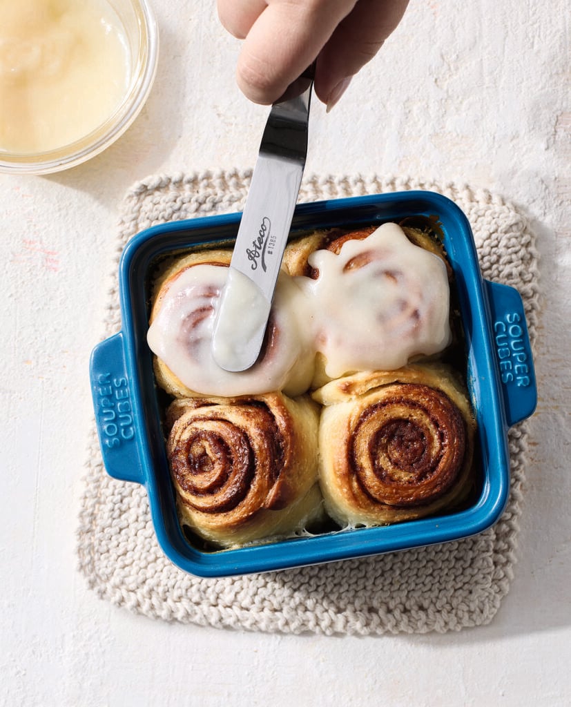 A hand spreads icing on cinnamon rolls in a blue square baking dish containing four rolls; two rolls are frosted. A small bowl of icing is in the upper left corner, and the dish rests on a knitted mat.