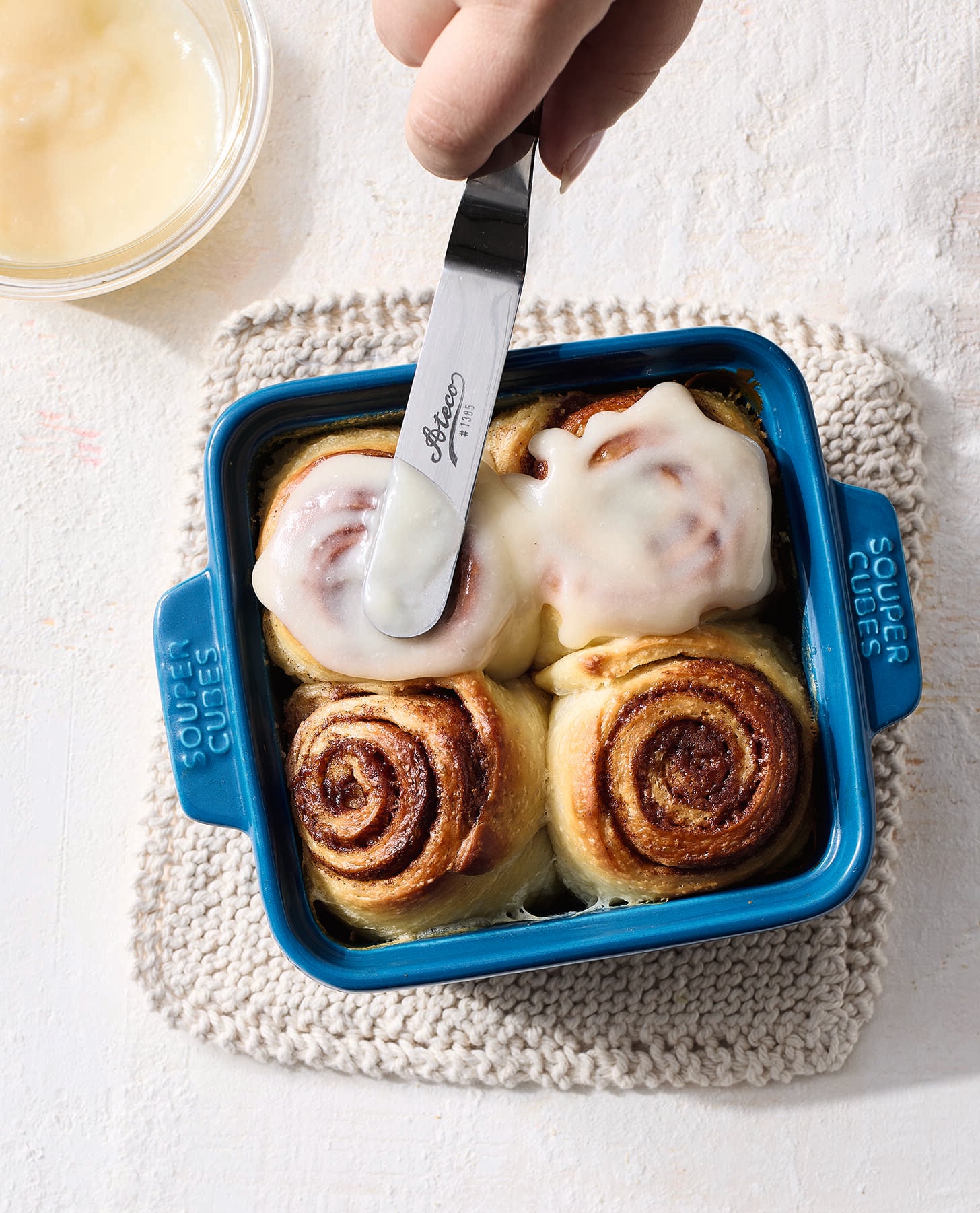 A hand spreads icing on cinnamon rolls in a blue square baking dish containing four rolls; two rolls are frosted. A small bowl of icing is in the upper left corner, and the dish rests on a knitted mat.