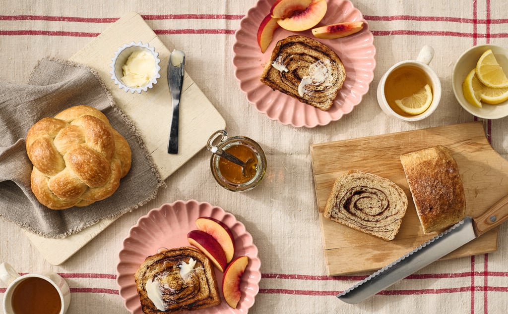 Breakfast scene with sliced cinnamon swirl bread, sliced peaches, braided bread, butter, honey jar with dipper, knives, and mugs of tea with lemon on a tablecloth with pink stripes.