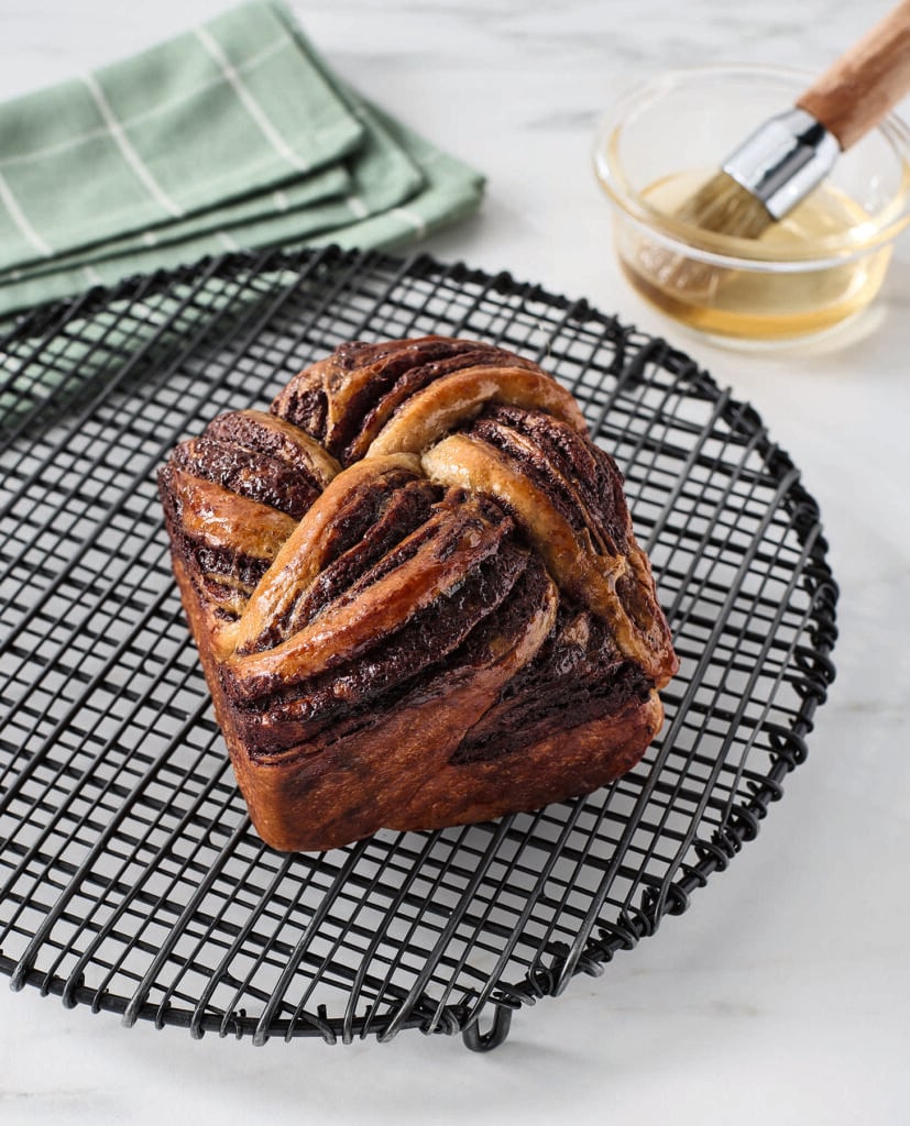 A loaf of twisted chocolate babka sits on a black cooling rack. In the background, a small bowl with a pastry brush and a folded green cloth are visible on a white surface.