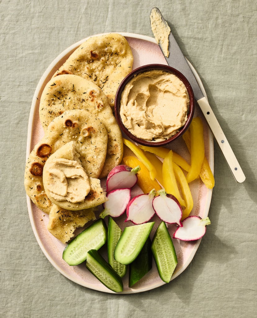 A plate with pieces of flatbread, a bowl of hummus with a knife, sliced yellow bell pepper, halved radishes, and sliced cucumber on a light green tablecloth.
