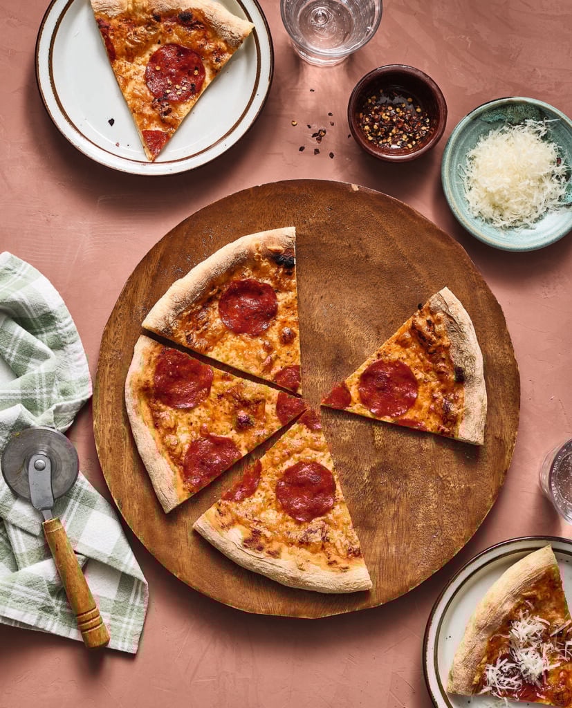 A pepperoni pizza on a wooden board with three slices missing; two of the missing slices are on small plates. Nearby are bowls of grated cheese, red pepper flakes, a pizza cutter, napkin, and glasses of water.