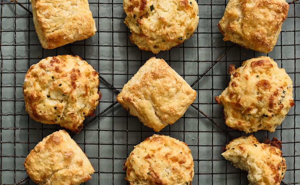 A variety of golden-brown biscuits, some round and some square, are cooling on a black wire rack placed over a green tiled surface.