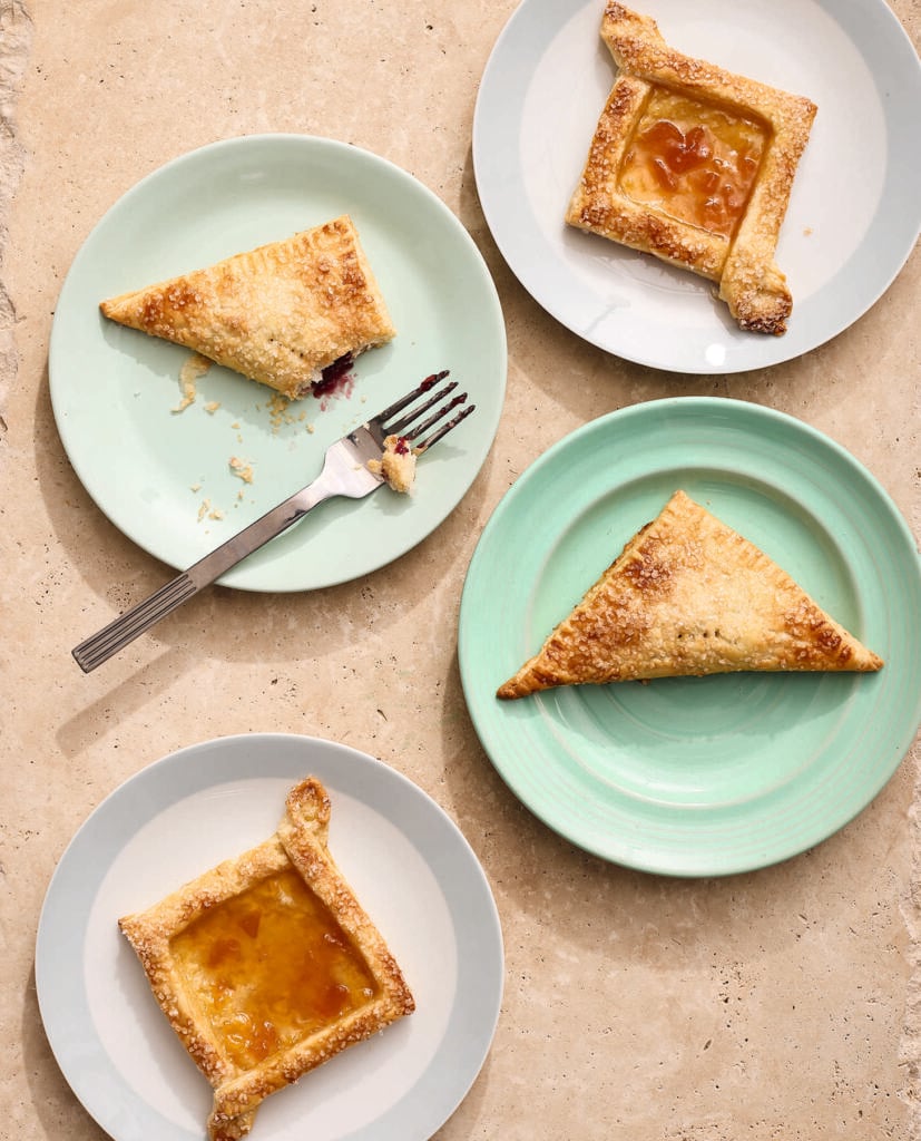 Four plates with baked pastries, two triangular and two square with jam filling, are arranged on a beige surface. One plate has a fork and a partially eaten pastry. The plates are light blue and pastel green.