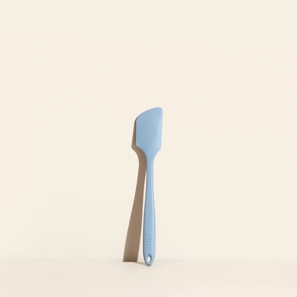 A light blue silicone spatula with a flat, angled edge is positioned vertically against a pale beige background. The handle has a small hole near the end for hanging.