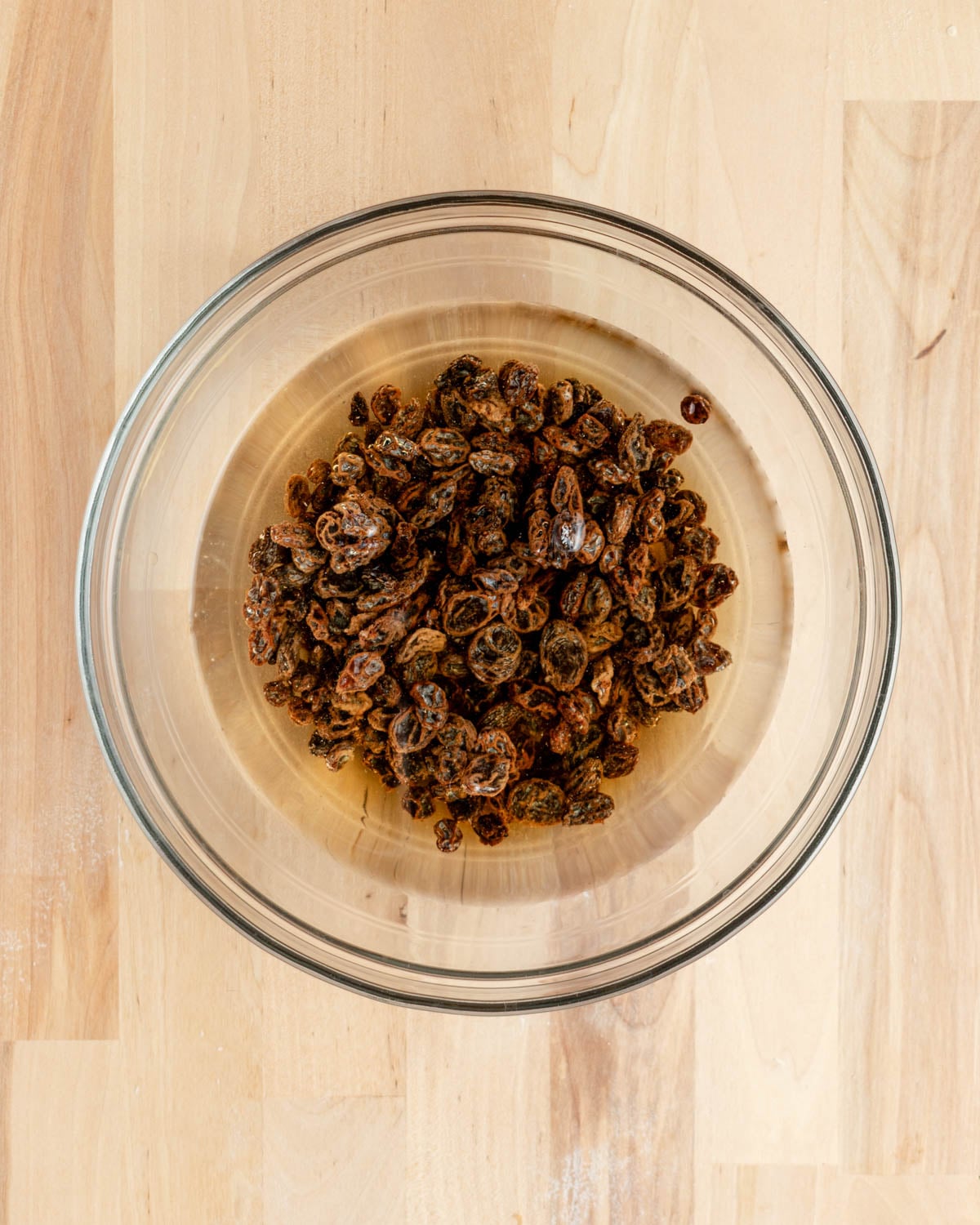 A clear glass bowl filled with dried raisins soaking in water sits on a light wooden surface, its contents visible from above.