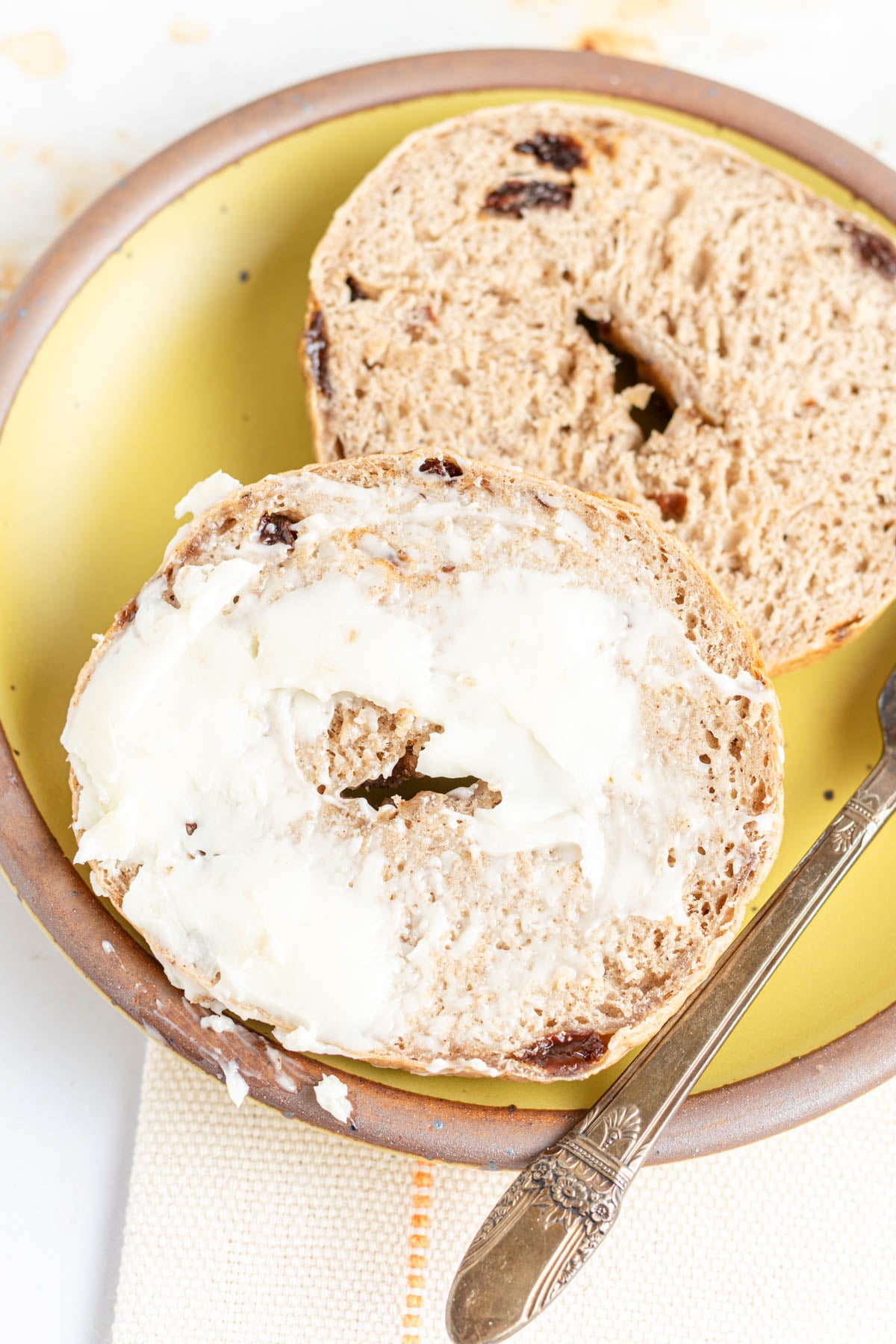 A sliced cinnamon raisin bagel sits on a yellow plate—one half spread with butter, the other plain. A butter knife rests nearby.