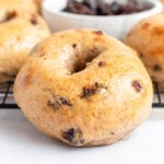 A close-up of a cinnamon raisin bagel with a shiny, golden-brown crust sits on a wire rack, while everything bagels and a bowl of raisins can be seen in the background.