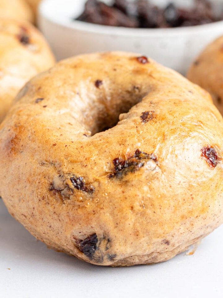 A close-up of a cinnamon raisin bagel with a shiny, golden-brown crust sits on a wire rack, while everything bagels and a bowl of raisins can be seen in the background.
